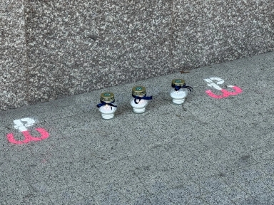 Trzy znicze ustawione na szarym chodniku przy ścianie z granitu, obok nich na chodniku namalowane symbole Polski Walczącej.