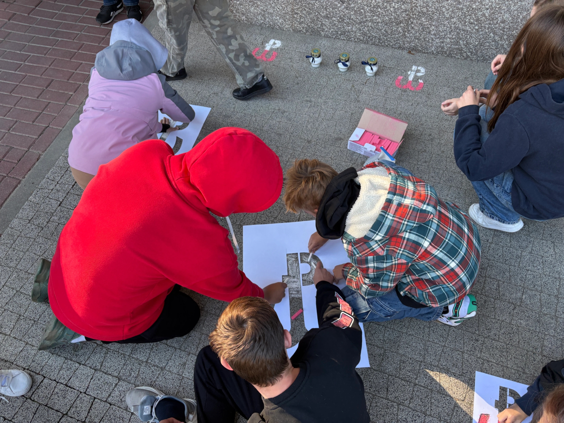 Grupa dzieci malujących litery na szablonach na chodniku podczas dnia.