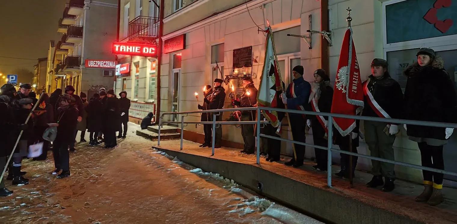 Nocna uroczystość na zaśnieżonym chodniku przed budynkiem. Po prawej stoi grupa osób na podwyższeniu z flagami i proporcami. Kilka osób trzyma zapalone pochodnie. Po lewej grupa uczestników stoi na chodniku. Czerwona iluminacja neonowego szyldu oświetla fasadę.