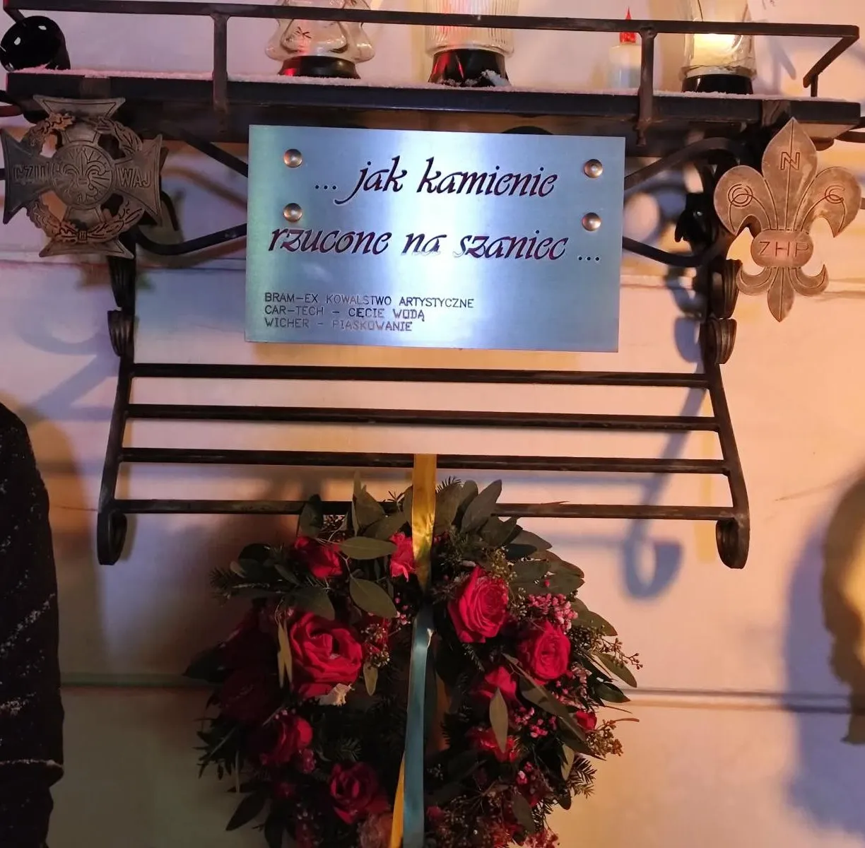 Metalowy stojak mocowany do jasnej ściany. Na środku przykręcona metalowa tabliczka z dwoma wkrętami. Po bokach metalowe emblematy: krzyż po lewej i lilijka harcerska po prawej. Pod stojakiem wisi wieniec z czerwonych róż i wstążkami.
