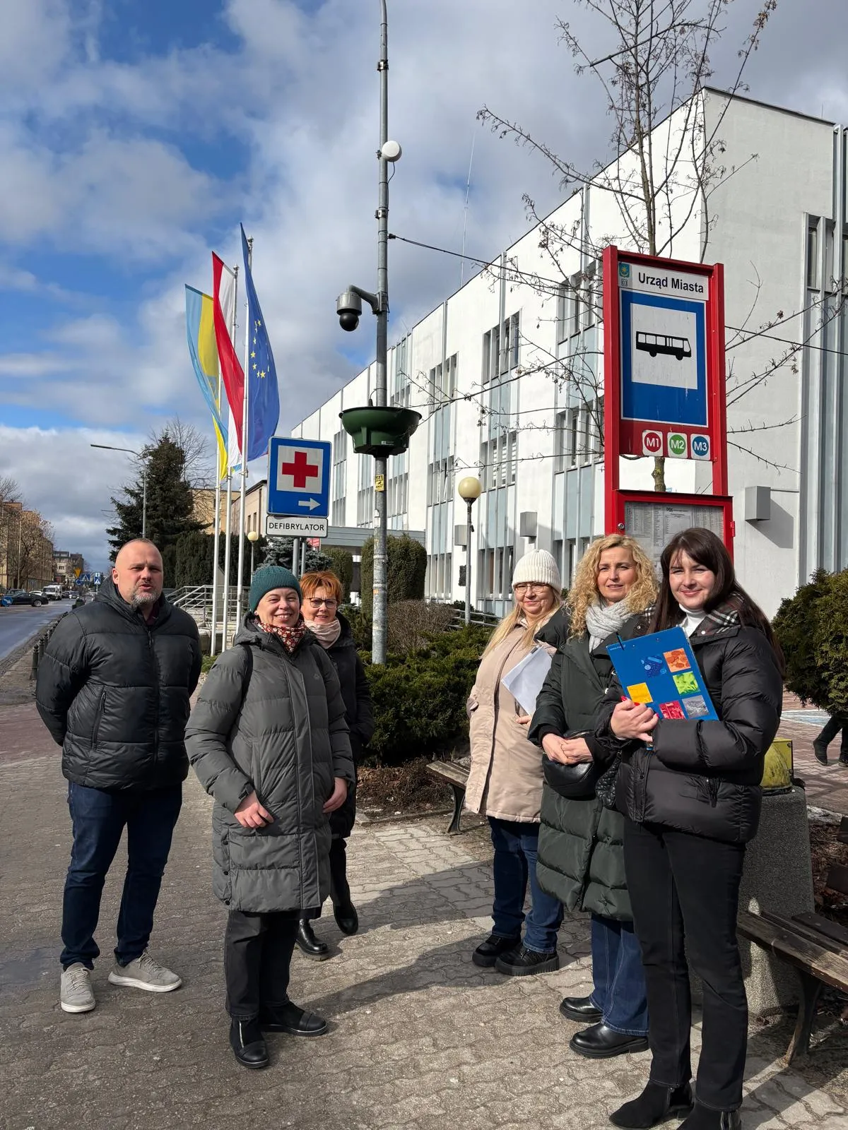 Sześć dorosłych osób stoi na chodniku przed białym budynkiem urzędu miasta. Osoby noszą zimowe kurtki i stoją przy przystanku autobusowym z niebieskim znakiem. Po lewej widać kilka pionowych flag, w tym flagę Unii Europejskiej. Na słupie widoczny jest znak z czerwonym krzyżem i napis o defibrylatorze. Niebo jest częściowo zachmurzone.