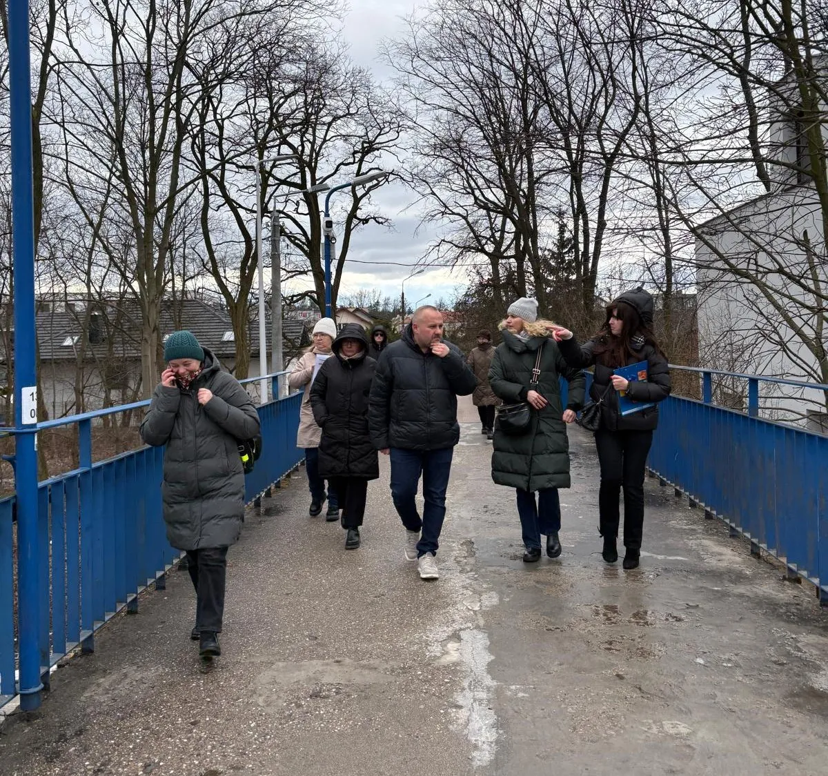 Siedem osób idzie w grupie po niebieskim kładce na zewnątrz. Ludzie mają zimowe płaszcze i czapki. Jest pochmurne niebo i nagie drzewa w tle.