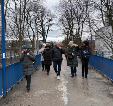 Siedem osób idzie w grupie po niebieskim kładce na zewnątrz. Ludzie mają zimowe płaszcze i czapki. Jest pochmurne niebo i nagie drzewa w tle.