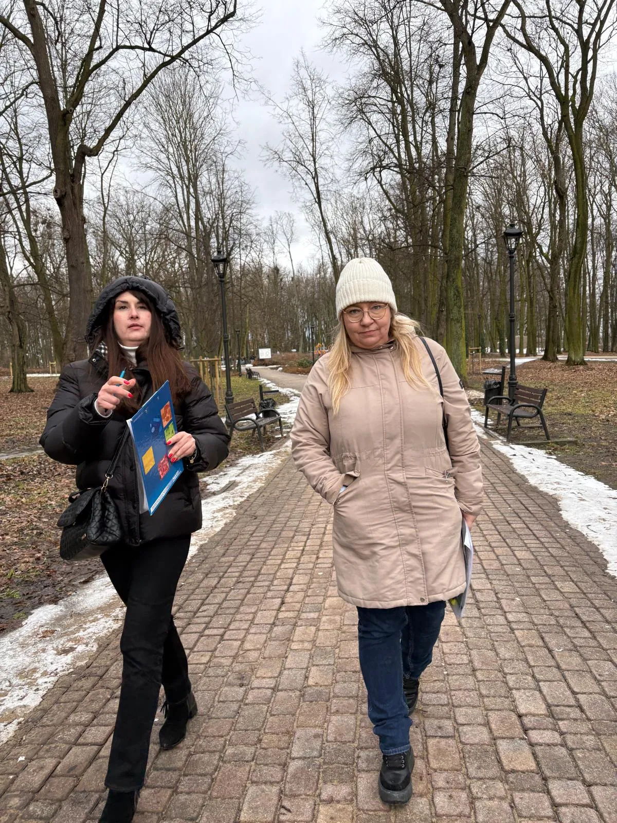 Dwie kobiety idą alejką w parku w zimowy, pochmurny dzień. Prawa ma jasny płaszcz i białą czapkę, trzyma dokumenty przy boku. Lewa ma czarną puchową kurtkę i trzyma kolorowy folder. W tle gołe drzewa, ławki i śniegowe resztki na trawie.