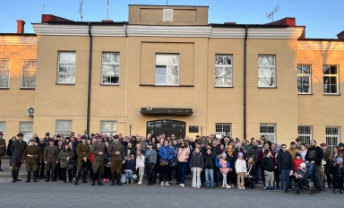 Duża grupa dorosłych i dzieci stoi przed dwukondygnacyjnym, beżowym budynkiem w ciągu dnia. Kilku mężczyzn w historycznych mundurach stoi z przodu po lewej stronie. W tłumie widać wózek dziecięcy, różne kurtki i okna budynku odbijające drzewa.