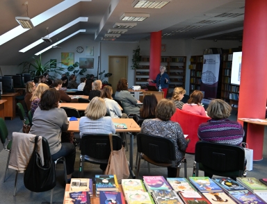 Wewnątrz biblioteki z dużymi świetlikami na skosnym suficie. Około dwudziestu dorosłych siedzi przy stolikach i słucha prowadzącej stojącej przy projektorze i banerze. Na pierwszym planie leżą ułożone książki, a wnętrze dzieli kilka czerwonych słupów i regałów z książkami.