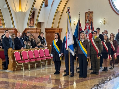 Wnętrze kościoła z grupą osób stojących podczas ceremonii. Trzy osoby na pierwszym planie trzymają sztandary. Po prawej stoi młodzież w szarfach biało-czerwonych i w mundurze. Po lewej rząd ławek z czerwonymi krzesłami i zebrani wierni. Marmurowa posadzka i łukowe sklepienie w tle.