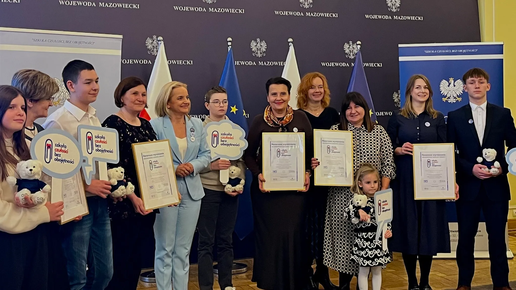 Grupa dwunastu osób stoi w pomieszczeniu z ciemnym tłem z napisem „WOJEWODA MAZOWIECKI” i flagami Polski oraz Unii Europejskiej. Kilka osób trzyma ramki z certyfikatami, pluszowe misie i tabliczki z napisem „szkoła czułości bez obojętności”. Wśród nich są dzieci i dorośli. Wszystkie osoby są ubrane formalnie lub półformalnie.