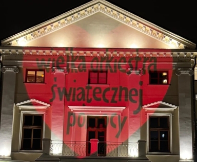 Budynek oświetlony nocą, na fasadzie biały klasyczny fronton i okna, z czerwonym sercem wyświetlonym na ścianie oraz czarnym napisem „wielka orkiestra świątecznej pomocy”.