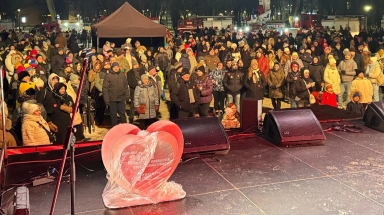 Duża grupa ludzi stojąca na zewnątrz w nocy przed sceną z nagłośnieniem i dużą dekoracją w kształcie serca, wszyscy ubrani w zimowe kurtki i czapki.