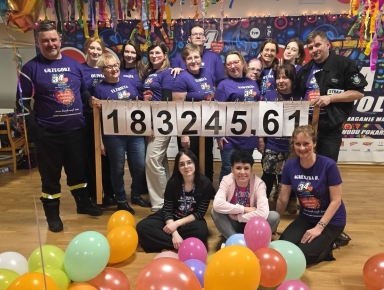 Grupa szesnastu osób wewnątrz pomieszczenia stoi i siedzi na podłodze. Większość osób nosi fioletowe koszulki z napisem i numerem 34. Trzy osoby trzymają tablicę z liczbą 183245,61. Na podłodze przed nimi znajduje się wiele kolorowych balonów. W tle kolorowa dekoracja ściany i wiszące kolorowe serpentyny.