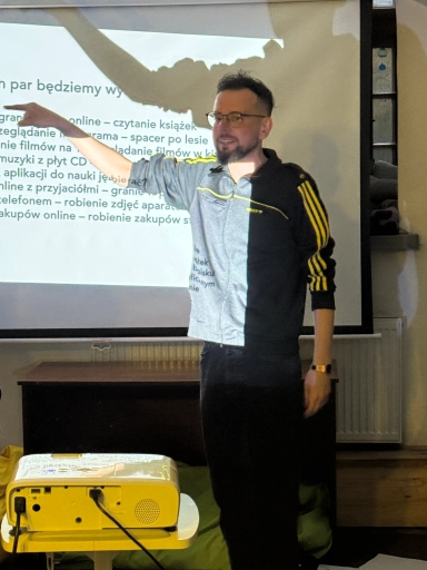 Mężczyzna w okularach stoi wewnątrz przy projekcji i wskazuje lewą ręką na biały ekran z tekstem; z przodu żółty projektor na stoliku, w tle gruby czarny mebel i jasna ściana.