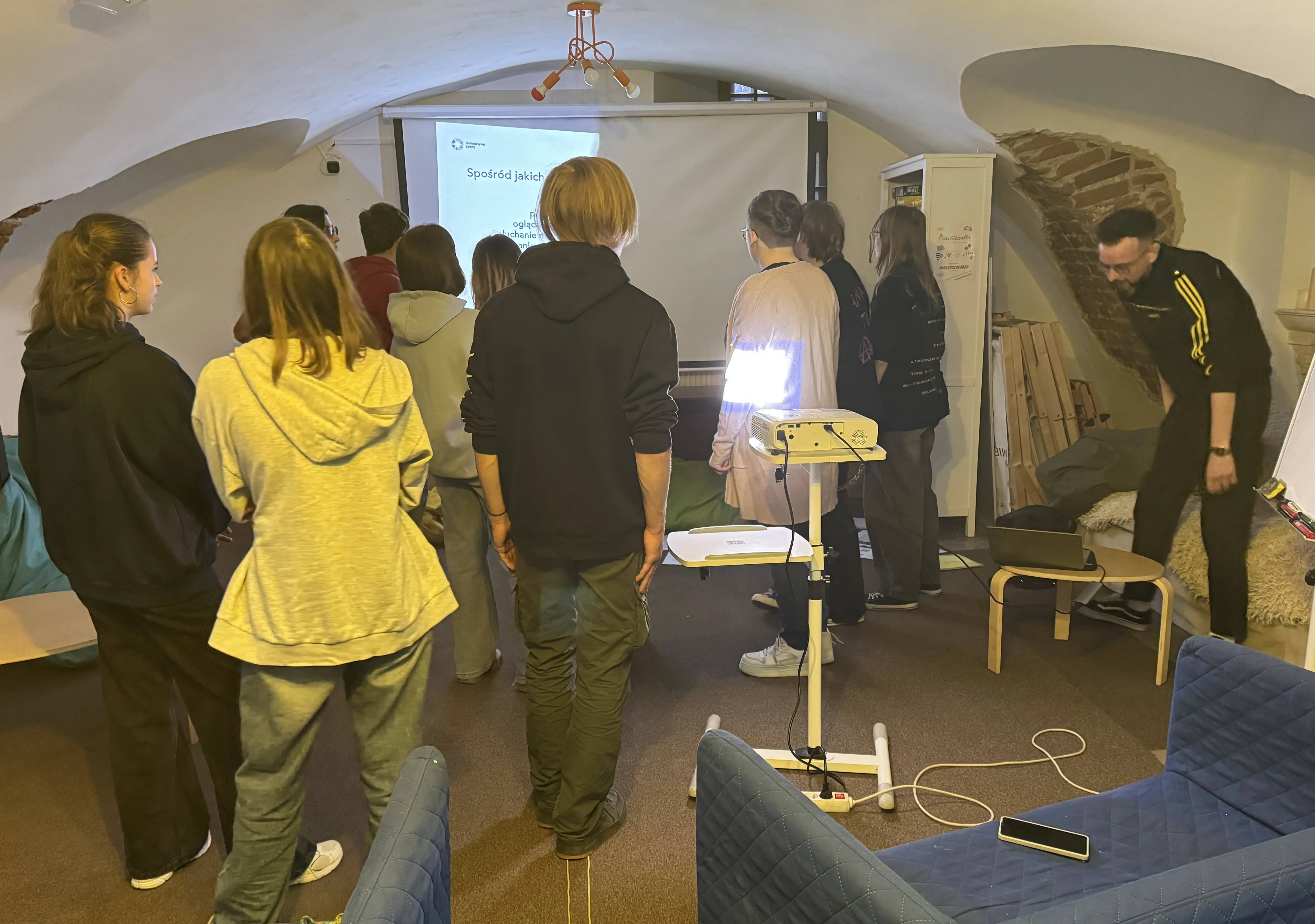 Grupa około dziesięciu młodych osób stoi tyłem do kamery i patrzy na biały ekran projekcyjny. W pomieszczeniu z zaokrąglonym sufitem i odsłoniętą cegłą po prawej stoi włączony projektor na statywie. Po prawej mężczyzna w ciemnym ubraniu schyla się nad niskim stolikiem, na pierwszym planie niebieska sofa i telefon.