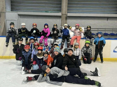 Grupa dzieci i trzech dorosłych na hali lodowiska, dzieci ubrane w kolorowe kombinezony i kaski, niektórzy siedzą na barierce, inni stoją lub leżą na lodzie, wnętrze z metalową siatką i ścianami w tle.