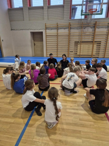 Dwadzieścioro dzieci i dwoje dorosłych siedzi w okręgu na podłodze drewnianej hali sportowej. W tle widać drabinki gimnastyczne, siatkę zabezpieczającą i kosz do koszykówki. Dzieci mają sportowe ubrania, sala jest oświetlona światłem dziennym.