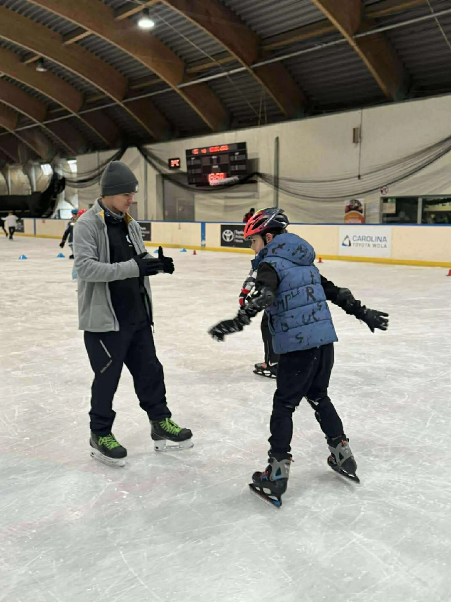 Dwóch mężczyzn jeździ na łyżwach po lodowisku krytym. Jeden ma szarą czapkę i kurtkę, drugi czerwoną kask i niebieską kamizelkę. W tle widać tarczę z zegarem i tablice reklamowe.