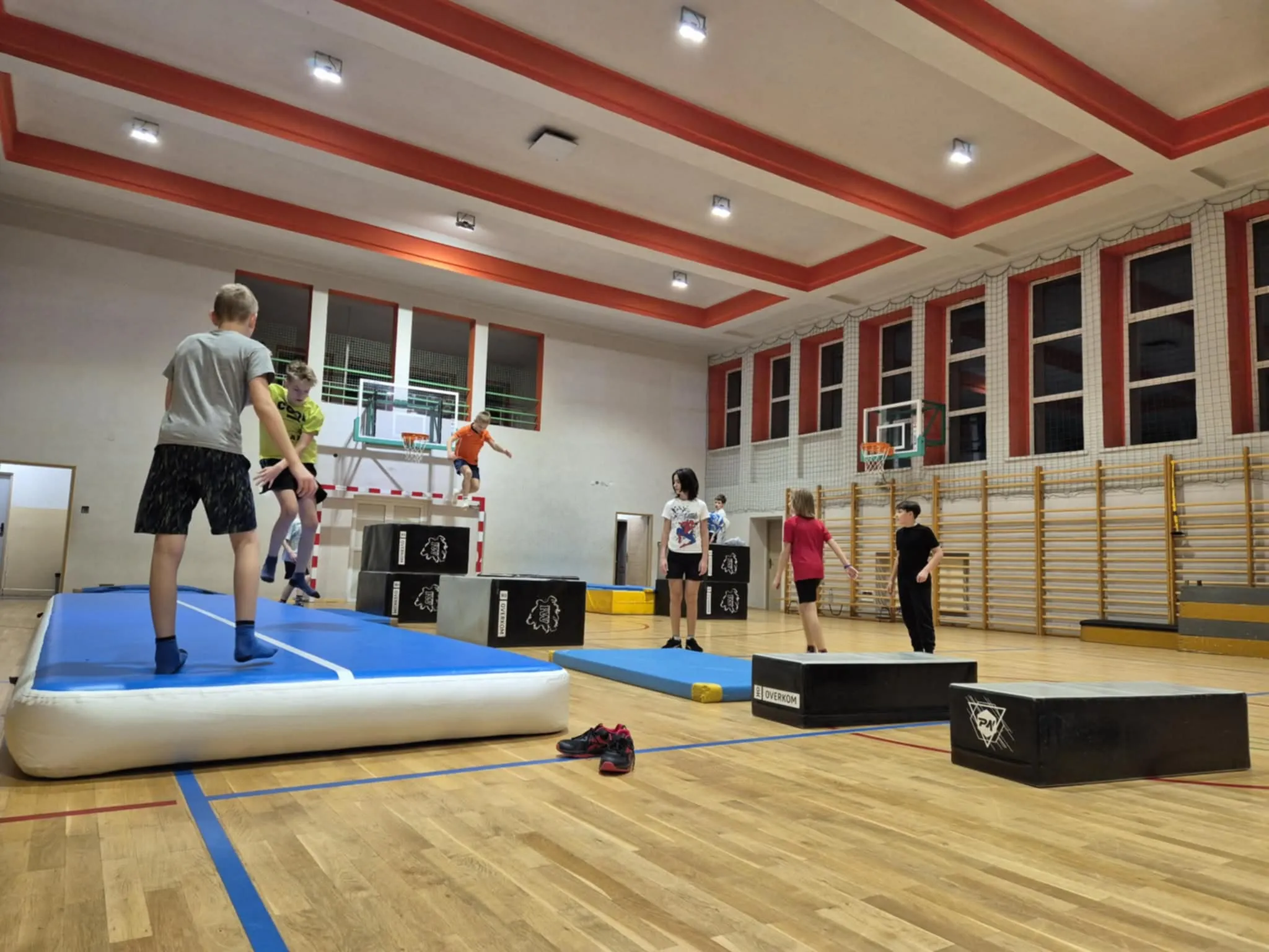 Wewnątrz hali sportowej dzieci ćwiczą na materacach i skrzyniach gimnastycznych rozstawionych na drewnianej podłodze. W tle widać kosze do koszykówki oraz ściankę do wspinaczki z drabinkami. Pomieszczenie jest jasne, z białymi ścianami i czerwonymi elementami na suficie oraz wokół okien.