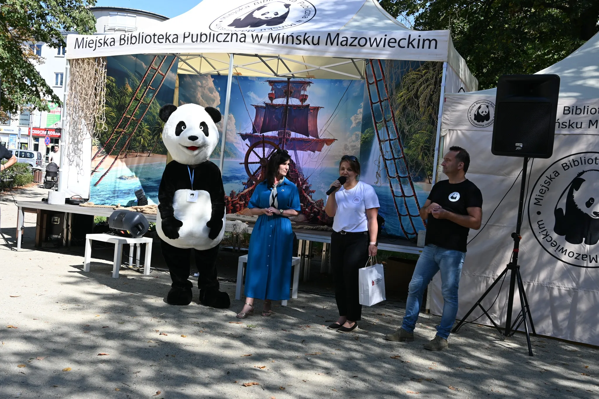Stoiska plenerowe Miejskiej Biblioteki Publicznej w Mińsku Mazowieckim z ludźmi, postacią w kostiumie pandy oraz dużym muralem z żaglowcem w tle, na zewnątrz w dzień.