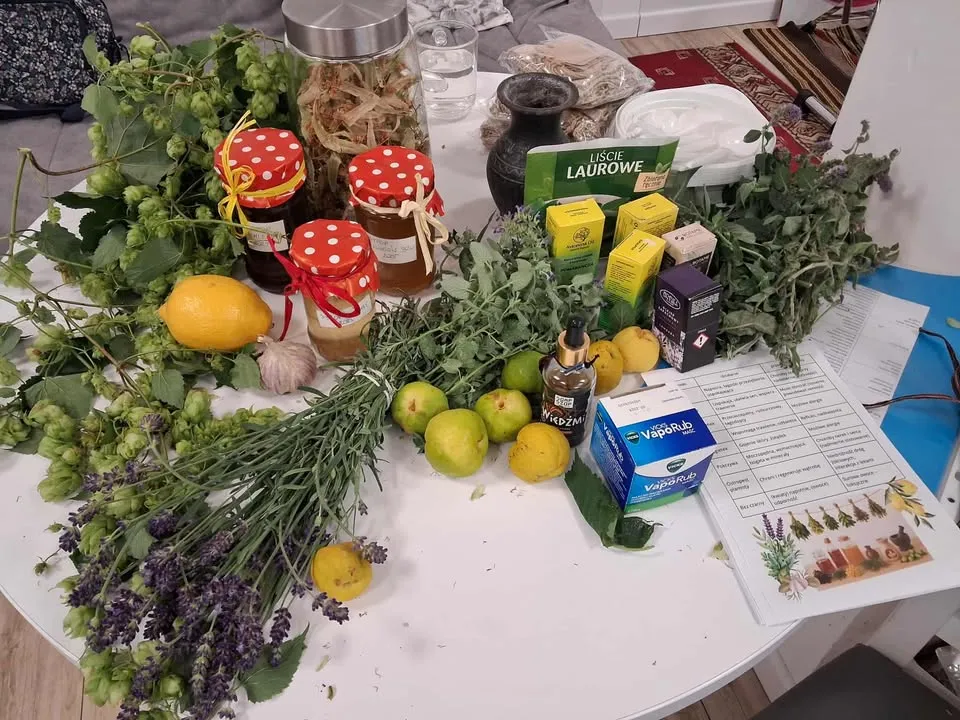 Stół z różnymi ziołami, owocami, przyprawami, miodami w słoikach, opakowaniami medykamentów oraz kartkami z informacjami.