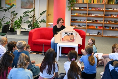 Kobieta prezentuje dzieciom opowieść obrazkową w bibliotece siedzi na czerwonej sofie na tle regału z książkami. Dzieci siedzą na dywanie i słuchają.