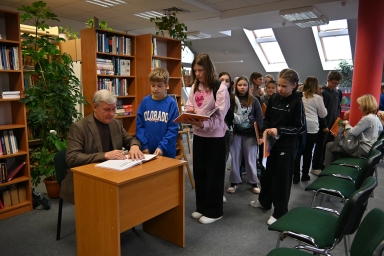 Mężczyzna siedzi przy wooden biurku w bibliotece i podpisuje książkę. Przed nim stoi kilkoro dzieci w kolejce trzymających książki. W tle widoczne regały z książkami, rośliny i duże okna dachowe. Po prawej stronie kilka zielonych krzeseł i dwie siedzące kobiety.
