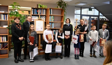 Grupa sześciu dzieci i czterech dorosłych stoi w bibliotece przed regałami z książkami. Dzieci trzymają białe torby i teczki. W tle jest plansza z napisem oraz rośliny doniczkowe. Pomieszczenie jest dobrze oświetlone sztucznym światłem.