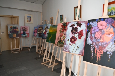 Wewnątrz korytarza stoi osiem sztalug w rzędzie przy białej ścianie. Na sztalugach wiszą kolorowe obrazy, głównie kwiatowe kompozycje i fragment twarzy w kwiatach. Po lewej widoczne drewniane drzwi. Na podłodze ciemnoszare płytki.