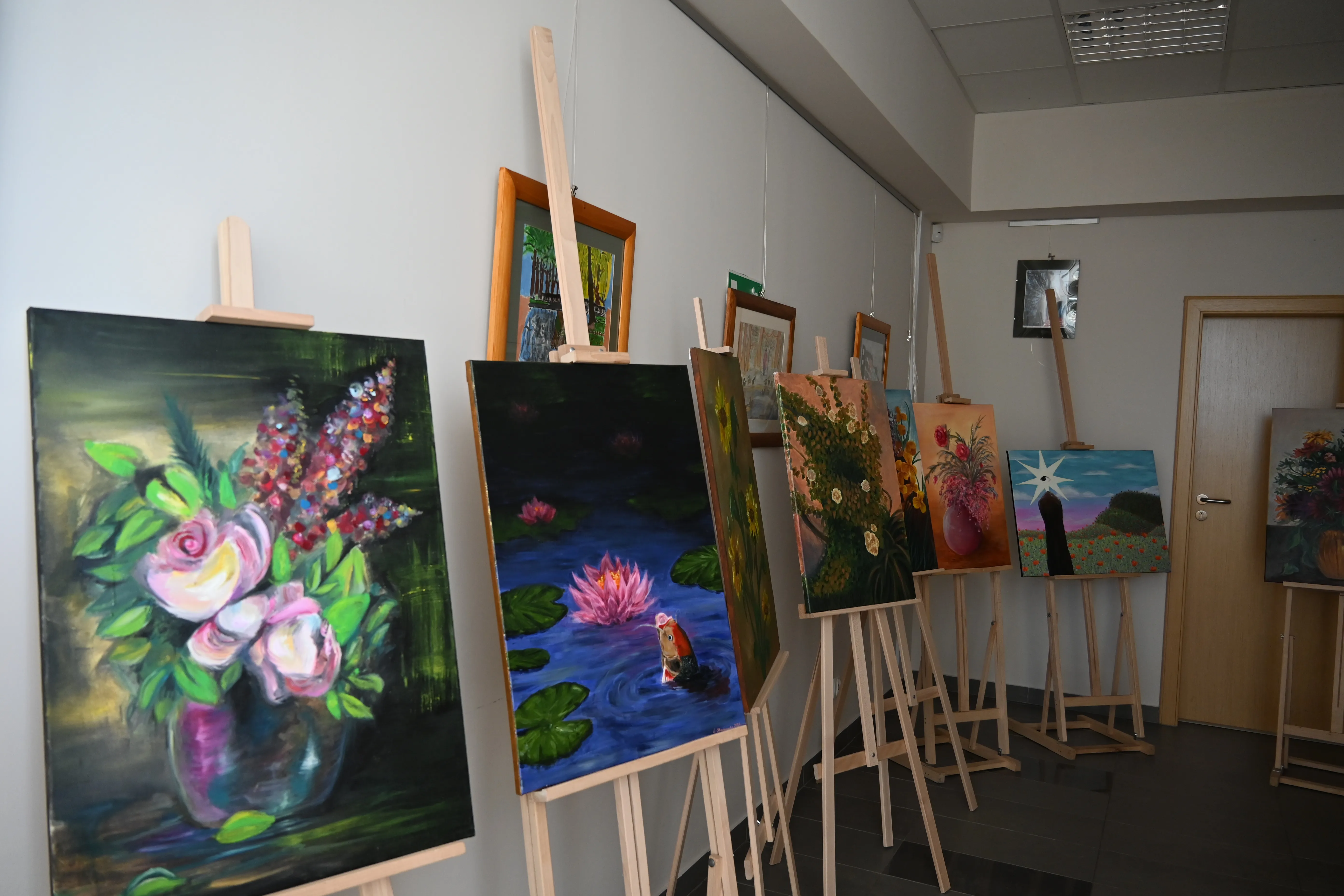 Kilka sztalug z obrazami ustawionych w rzędzie przy jasnoszarej ścianie w wnętrzu. Obrazy przedstawiają kwiaty, lilie wodne i kolorowe kompozycje na ciemnych i jasnych tłach. Po prawej drzwi drewniane, nad sufitem panele świetlne.