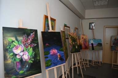 Kilka sztalug z obrazami ustawionych w rzędzie przy jasnoszarej ścianie w wnętrzu. Obrazy przedstawiają kwiaty, lilie wodne i kolorowe kompozycje na ciemnych i jasnych tłach. Po prawej drzwi drewniane, nad sufitem panele świetlne.