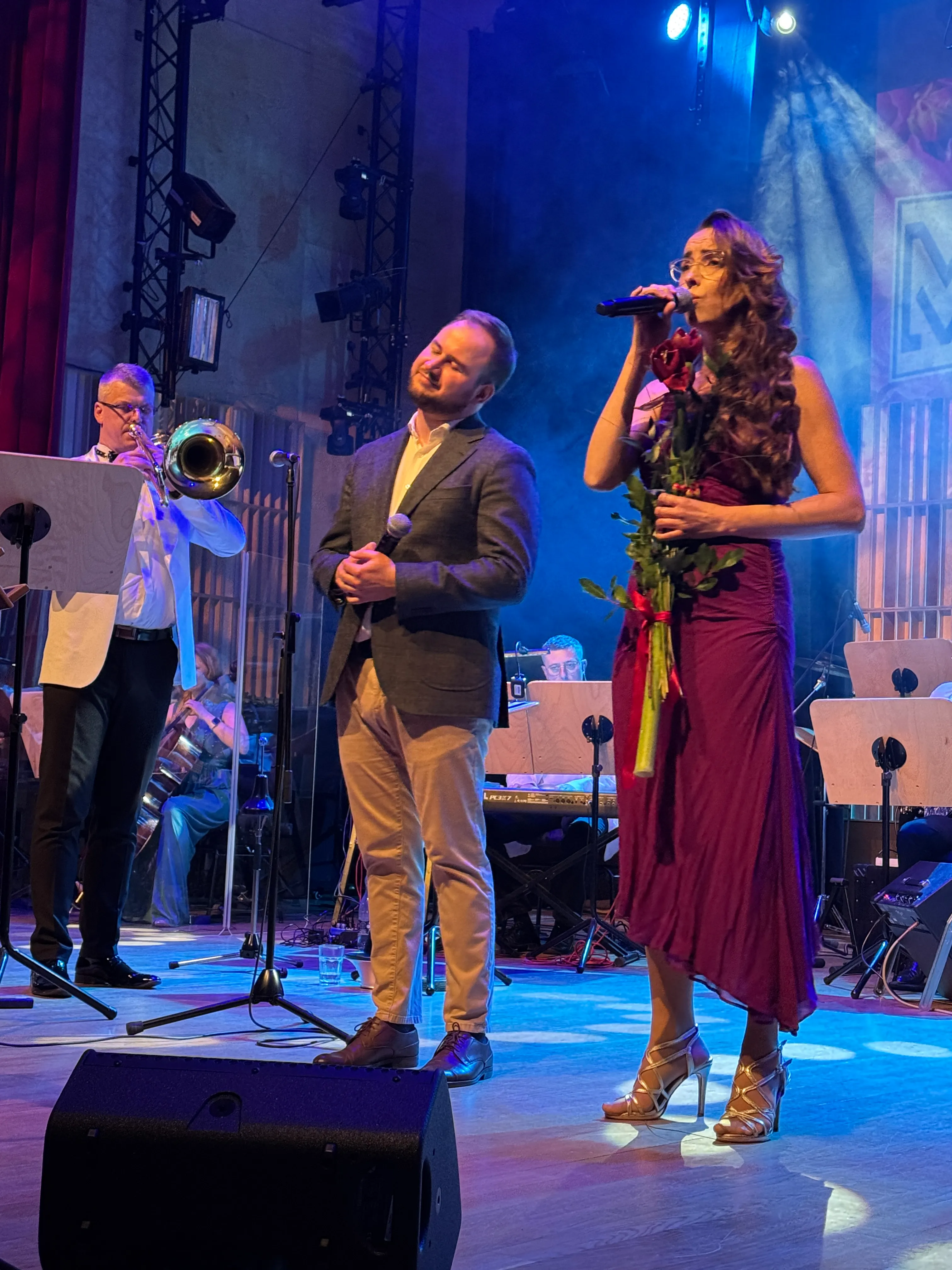 Na scenie stoi trzech wykonawców pod niebieskim oświetleniem. Kobieta w długiej fioletowej sukni i złotych szpilkach śpiewa do mikrofonu i trzyma bukiet czerwonych róż. Mężczyzna w marynarce obok trzyma mikrofon i stoi z zamkniętymi oczami, a po lewej muzyk w białej marynarce gra na trąbce; w tle siedzi orkiestra.