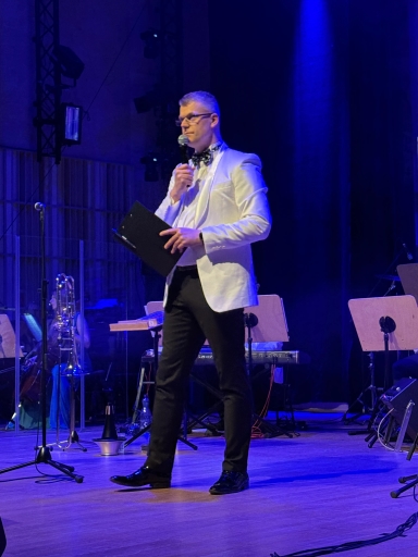 Mężczyzna w białej marynarce i czarnych spodniach stoi na scenie. Trzyma mikrofon przy ustach i czarną teczkę. Scena jest oświetlona niebieskim światłem, w tle stoją stojaki z nutami i instrumenty orkiestry.