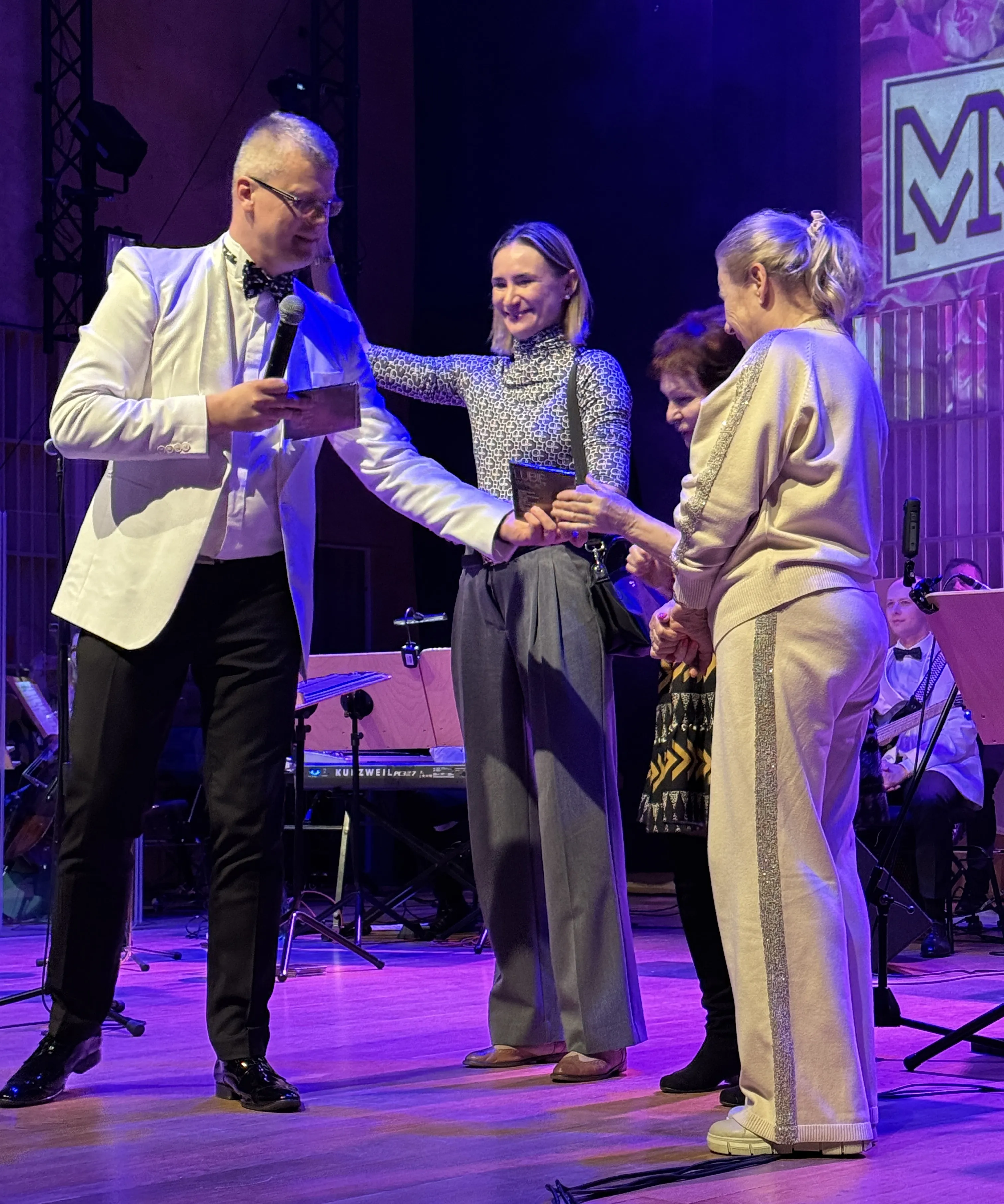 Mężczyzna w białej marynarce z mikrofonem podaje kartkę trzem kobietom na scenie. Kobieta w środku uśmiecha się i wyciąga dłoń, starsza kobieta po prawej stoi bokiem w jasnym dresie z brokatowym paskiem, trzecia kobieta częściowo zasłonięta w ciemnej sukience. Scena jest wewnątrz, oświetlona fioletowymi reflektorami, w tle instrumenty i muzycy.