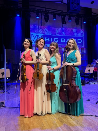 Cztery kobiety stoją na scenie obok siebie. Każda trzyma instrument smyczkowy: trzy mniejsze skrzypce/altówki i jedną kontrabas. Kobiety noszą długie suknie w kolorach koralowym, kremowym, miętowym i morskim. Scena ma drewnianą podłogę i niebieskie oświetlenie z tylnym panelem scenicznym.