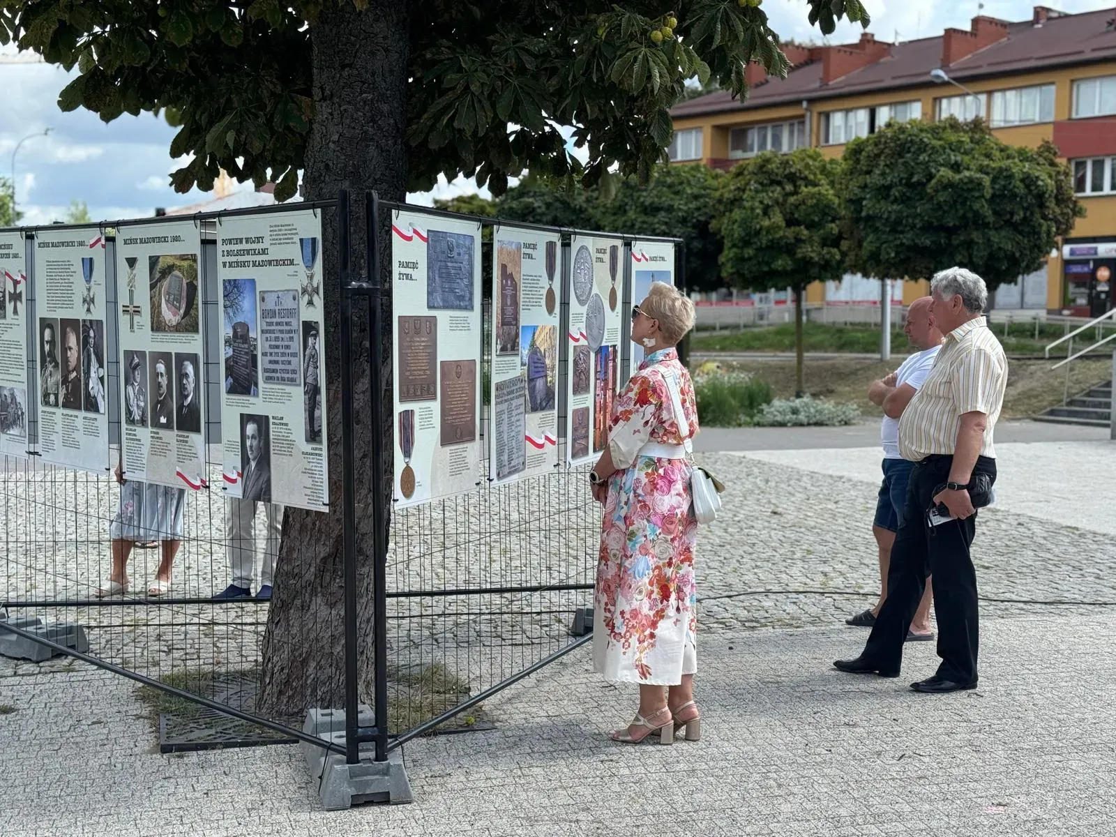 Na zewnątrz. Trzy osoby stoją przy planszach z wystawą historyczną umieszczoną wokół drzewa na placu z kostki brukowej, w tle drzewa i budynki mieszkalne.