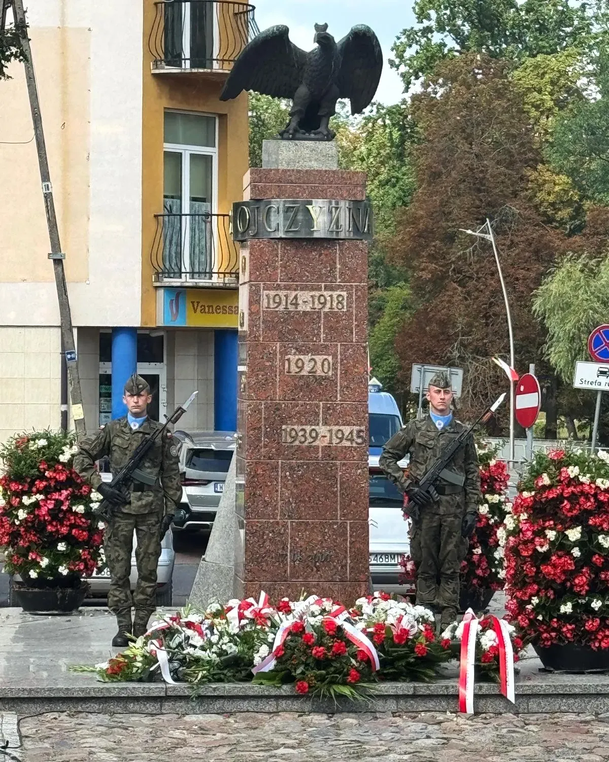 Na zewnątrz. Pomnik z orłem na szczycie i napisami upamiętniającymi, otoczony kwiatami. Obok stoi dwóch żołnieżyw mundurach wojskowych z bronią.