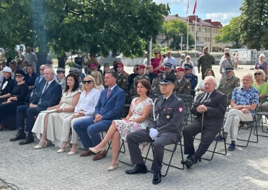 Na zawnątrz. Słoneczny dzień. Grupa około trzydziestu osób siedzi na krzesłach. Wśród nich osoby w mundurach wojskowych.