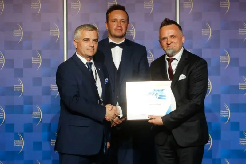 Trzech mężczyzn w garniturach stoi na tle niebieskiego baneru, dwóch z nich ściska dłonie, a jeden trzyma dyplom.