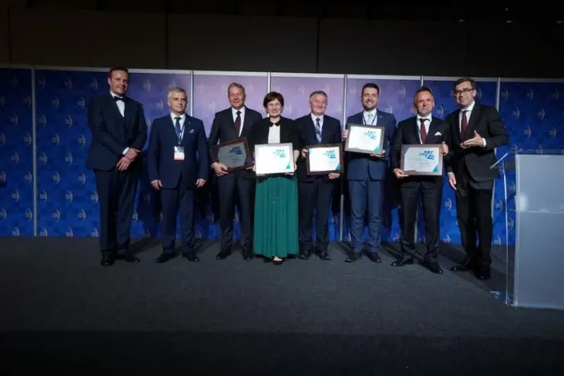 Siedem osób w formalnych strojach stoi na scenie, a cztery z nich trzymają dyplomy lub nagrody.