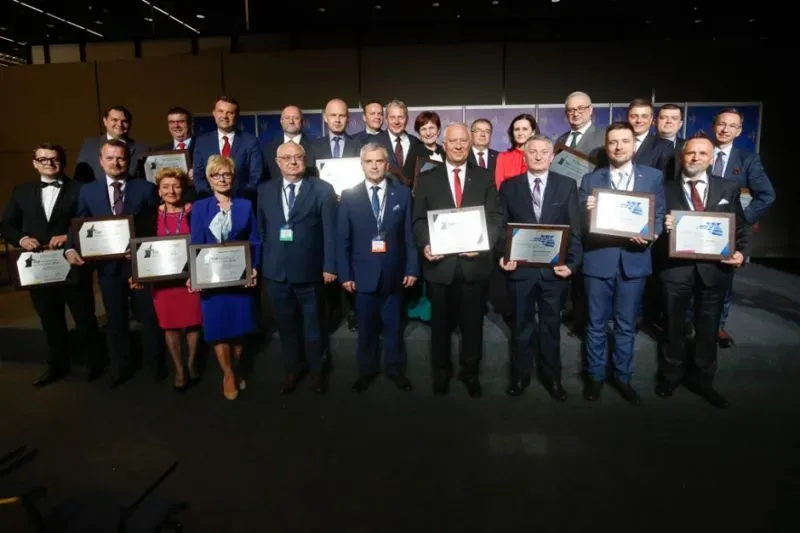 Dwudziestu pięciu ubranych formalnie dorosłych ludzi stoi w grupie, z których dziesięć osób trzyma w rękach nagrody lub dyplomy.