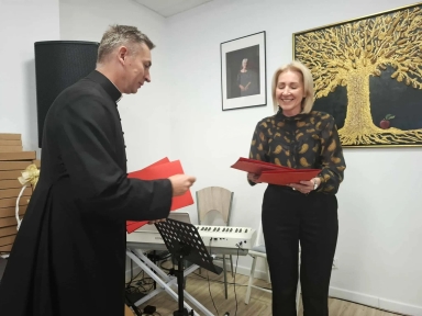 Jasne pomieszczenie. Ksiądz w czarnym stroju duchownym i uśmiechnięta kobieta w czarnej bluzce w pomarańczowy wzór wymieniają się czerwonymi folderami.