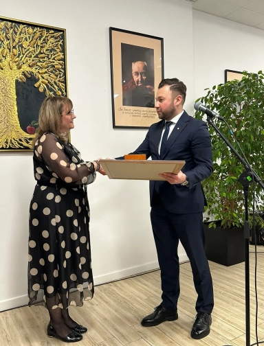 Jasne pomieszczenie. Mężczyzna w granatowym garniturze przekazuje kobiecie w czarnej sukience w beżowe grochy ramkę z wyróżnieniem, stojąc na jasnej podłodze. W tle na ścianie wiszą dwa obrazy, po prawej stronie stoi mikrofon na statywie i wysoka zielona roślina w doniczce.