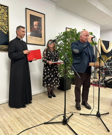 Jasne pomieszczenie. Trzy osoby stoją na jasnej podłodze. Mężczyzna po prawej mówi do mikrofonu, trzymając opakowanie w rękach. Po lewej stronie stoi ksiądz w czarnej sutannie z czerwoną teczką. Między nimi stoi kobieta w czarnej sukni w beżowe grochy, trzyma dokumenty. Za nimi wysoka roślina doniczkowa, na ścianie wiszą dwa obrazy, w tym portret starszego mężczyzny.