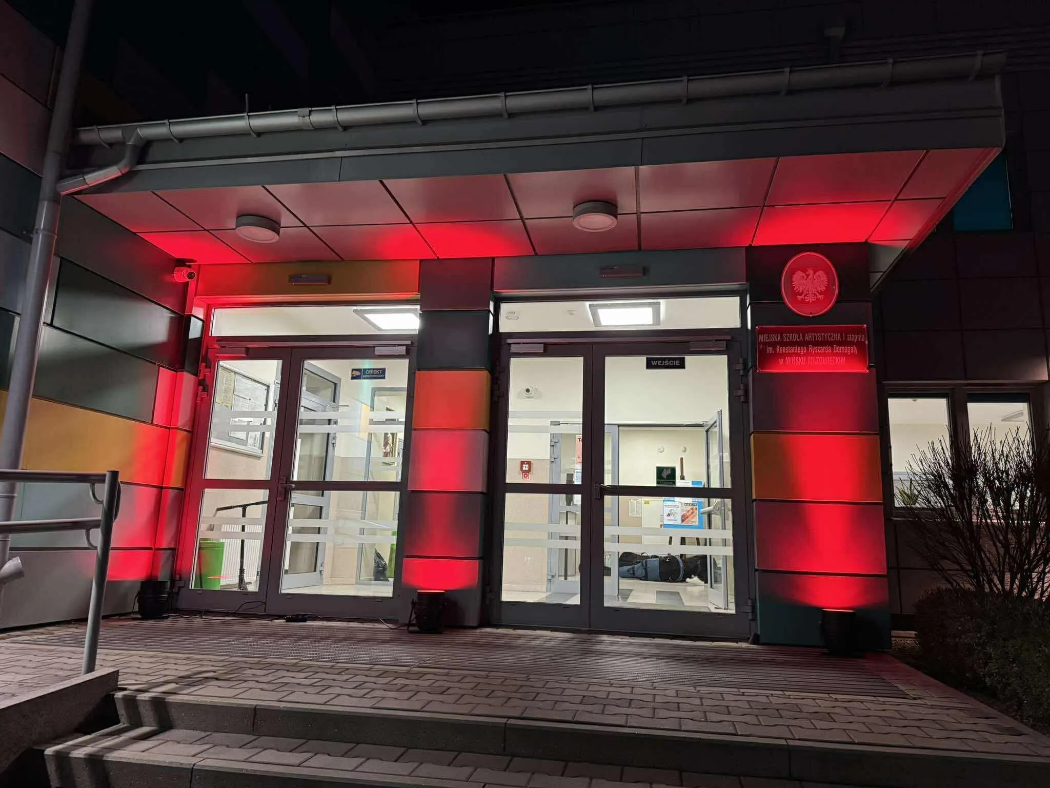 Wejście do budynku w nocy, oświetlone na czerwono wokół drzwi. Przed budynkiem schody i poręcz, po prawej stronie krzewy. Wnętrze widoczne przez przeszklone drzwi dobrze oświetlone.