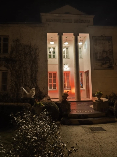 Wejście do budynku muzeum oświetlone nocą, dwie kolumny przy wejściu, po bokach donice z roślinami, przy ścianie rzeźba, na elewacji pnącza.