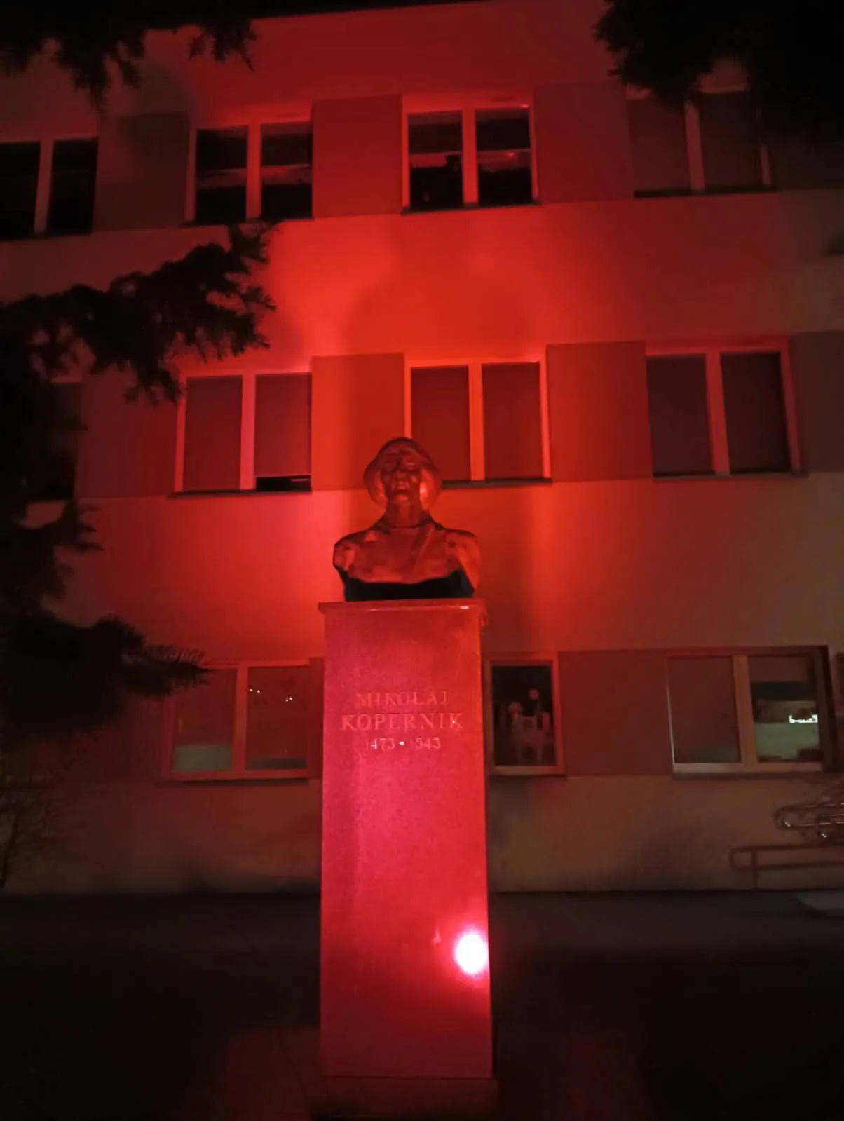 Popiersie Mikołaja Kopernika na cokole oświetlone czerwonym światłem, w tle budynek z oknami, noc, częściowo widoczne gałęzie drzew.