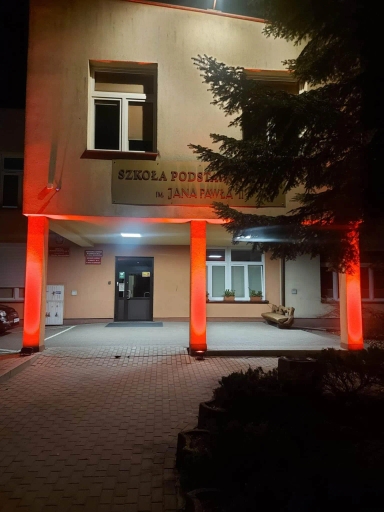 Budynek szkoły oświetlony nocą czerwonym światłem na czterech filarach pod wejściem. Drzwi wejściowe z ciemnym szkłem, po prawej stronie ławka i dwa okna z kwiatami. Z prawej strony zdjęcia widoczne gałęzie dużego drzewa. Przed wejściem brukowany podjazd.