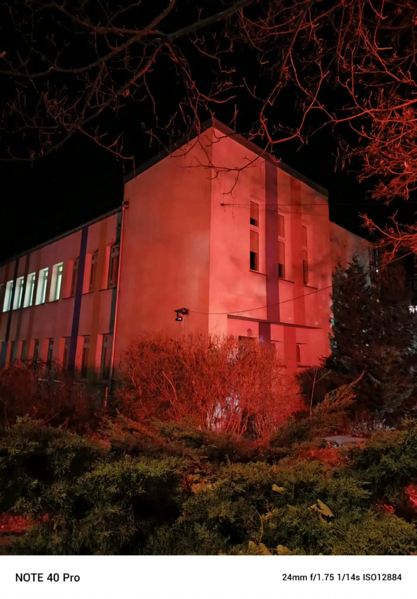 Dwupiętrowy budynek nocą oświetlony czerwonym światłem. Przed budynkiem znajdują się krzewy i drzewa bez liści, widoczne gałęzie na czarnym niebie. Część okien jest oświetlona światłem białym.