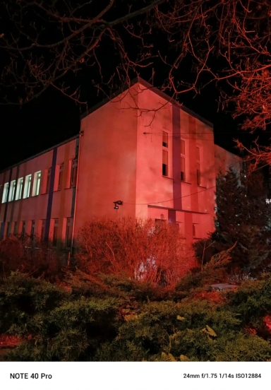 Dwupiętrowy budynek nocą oświetlony czerwonym światłem. Przed budynkiem znajdują się krzewy i drzewa bez liści, widoczne gałęzie na czarnym niebie. Część okien jest oświetlona światłem białym.