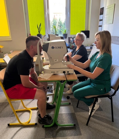 Mężczyzna siedzi naprzeciw kobiety w zielonym ubraniu medycznym, która obsługuje urządzenie okulistyczne, a druga kobieta siedzi obok przy biurku w jasnym pomieszczeniu z oknami i roślinami.