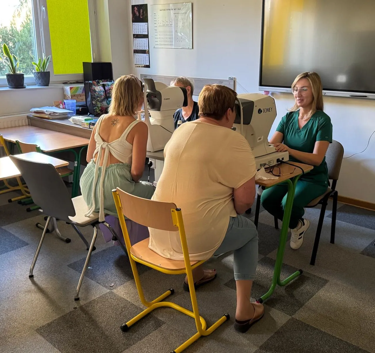 Dwie kobiety siedzą naprzeciw siebie w gabinecie, gdzie lekarz w zielonym ubraniu wykonuje badanie wzroku za pomocą specjalistycznych aparatów.